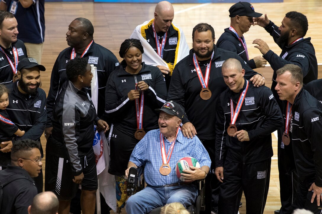 A veteran in a wheelchair sits surrounded by Team Army members wearing bronze medals and black uniforms in a gym.