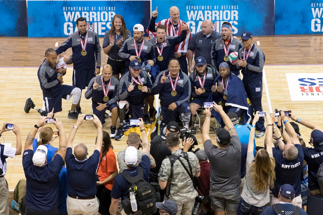 Team Navy pose wearing gold medals and holding up their index fingers as crowd members photograph them.