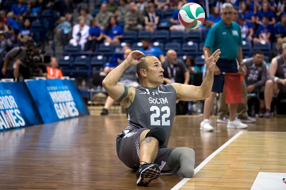 A soldier sitting on a gym floor stares up at a volleyball suspended in midair as he executes a serve.