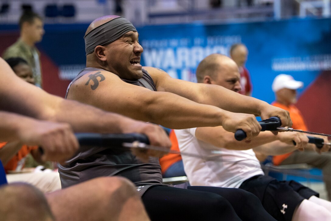 A sailor winces while pulling the handle of a rowing machine, alongside fellow competitors doing the same in a gym.