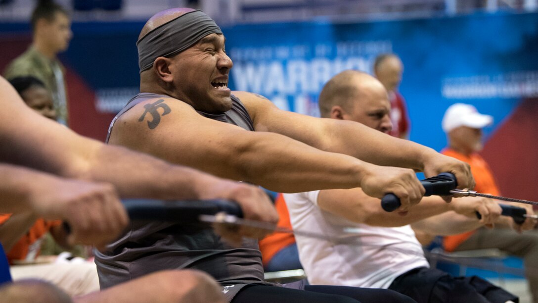 A sailor winces while pulling the handle of a rowing machine, alongside fellow competitors doing the same in a gym.