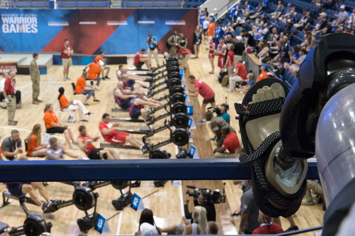 A prosthetic foot rests on a blue metal railing in stands overlooking a gym floor where athletes compete on rowing machines.