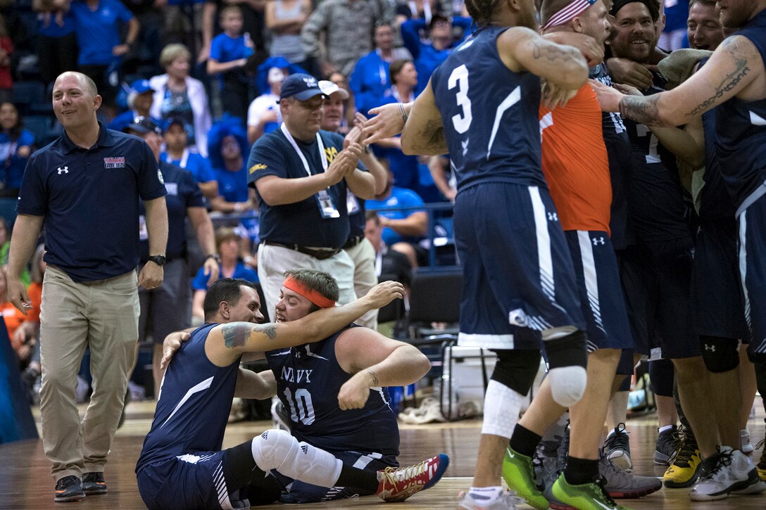 Two athletes seated on a gym floor hug as others huddle nearby, smiling and talking.