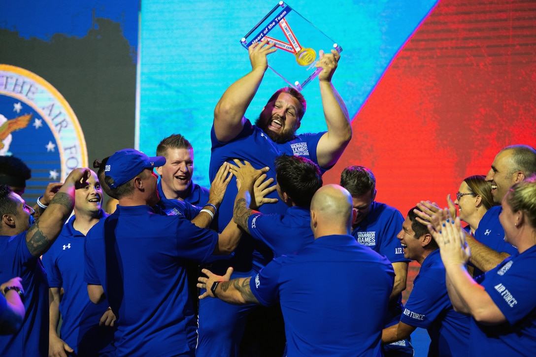 Athletes in blue uniforms hoist a teammate, who lifts a medal enclosed in glass over his head.