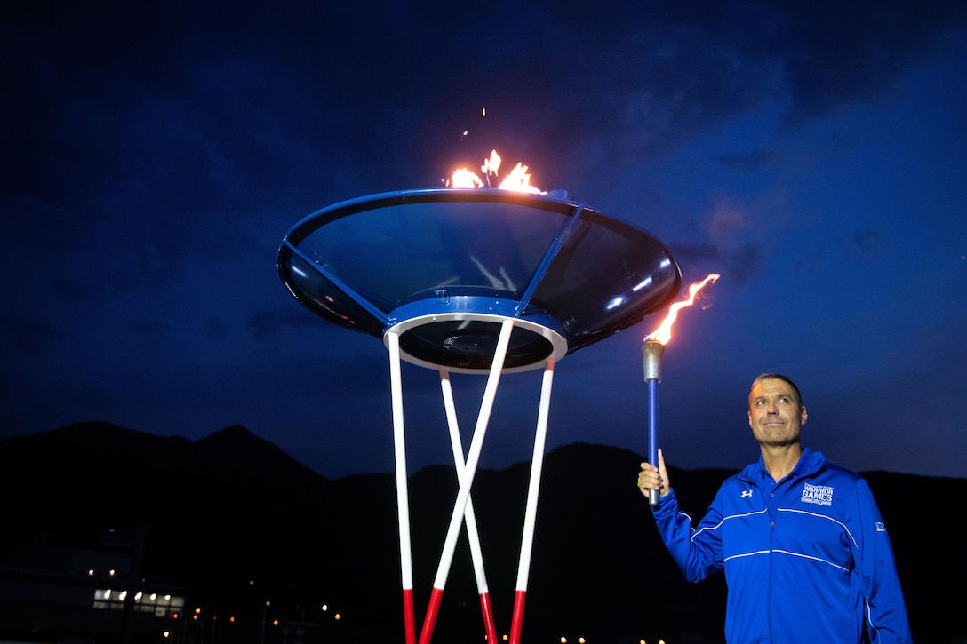 An airman holds a lit torch while standing beside a raised cauldron with a fire in it.