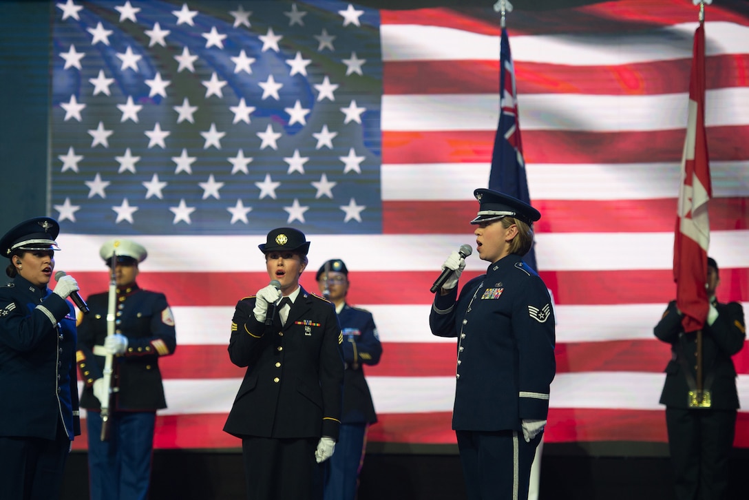 Service members sing on a stage, with an American flag on a screen in the background.