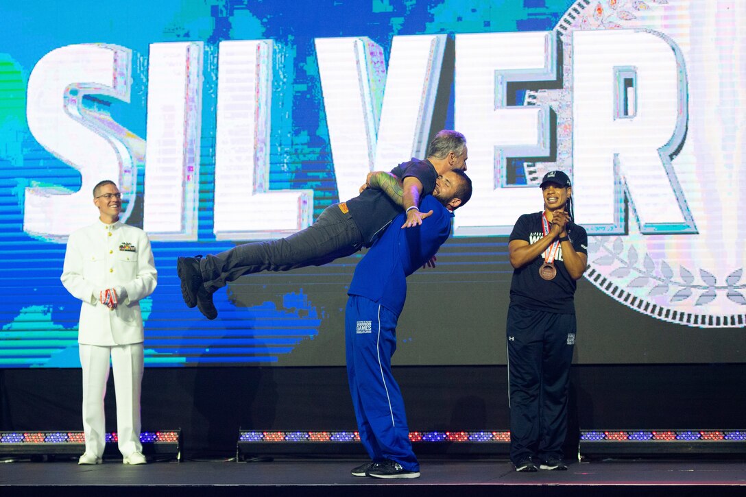 An athlete lifts comedian Jon Stewart off his feet, as two others watch and applaud.