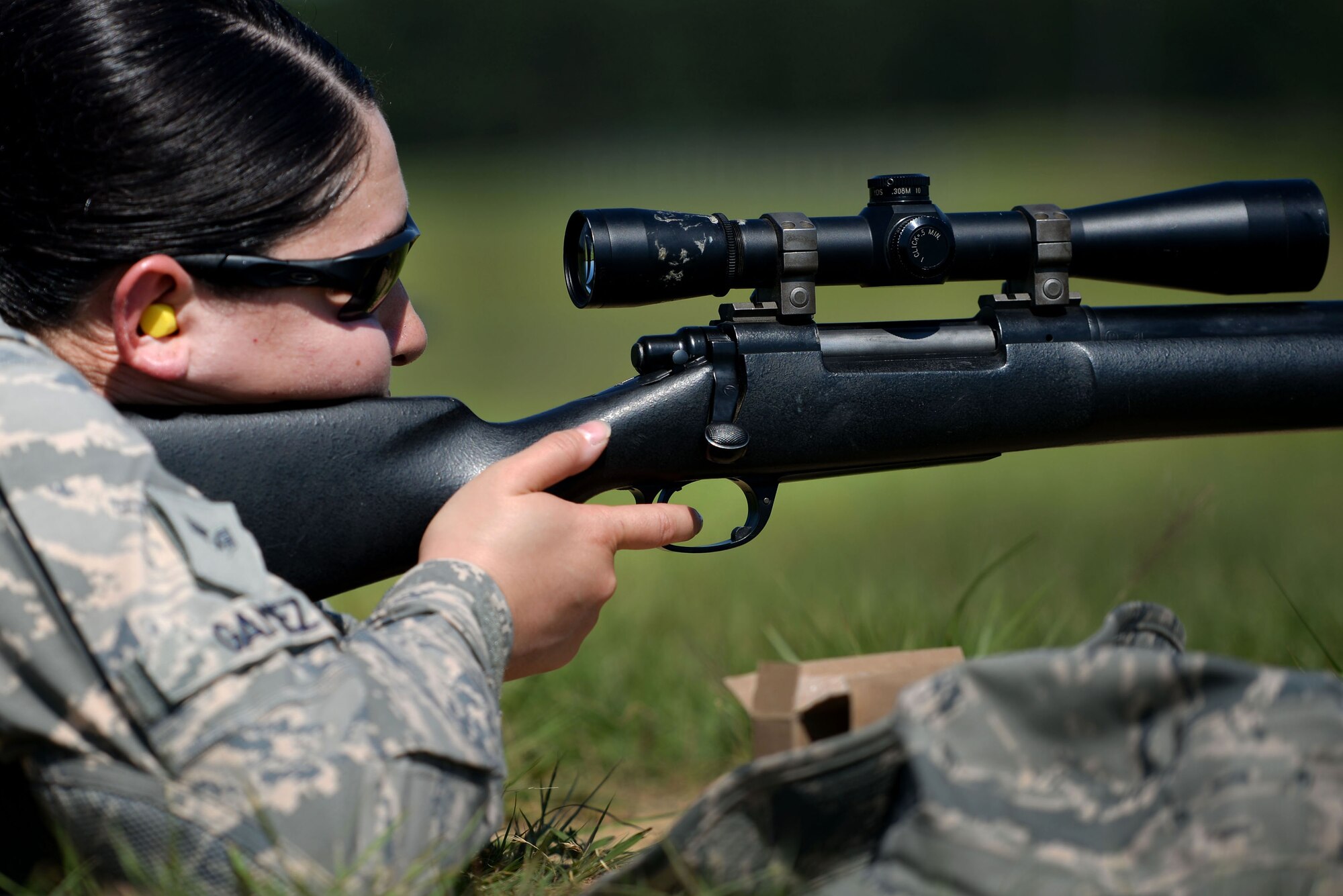 A woman in the Airman Battle Uniform and sunglasses peers down the scope of an M24 sniper weapon system.