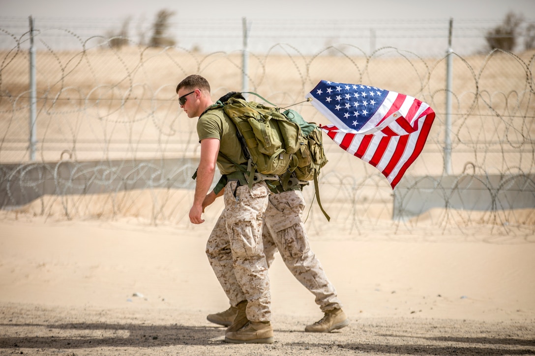 Two Marines carry an American flag as they hike by a barbed wire fence in desert terrain.