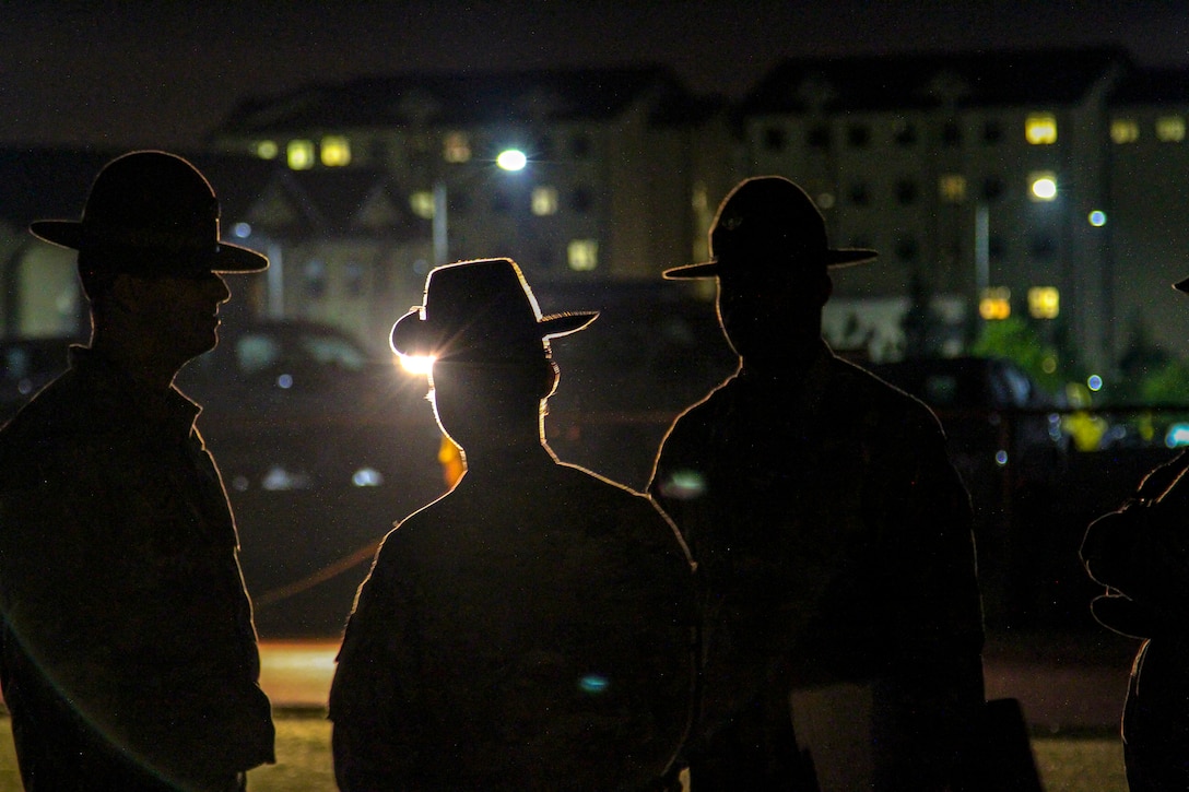 Three sergeants, shown in silhouette, look across a field toward a building at night.