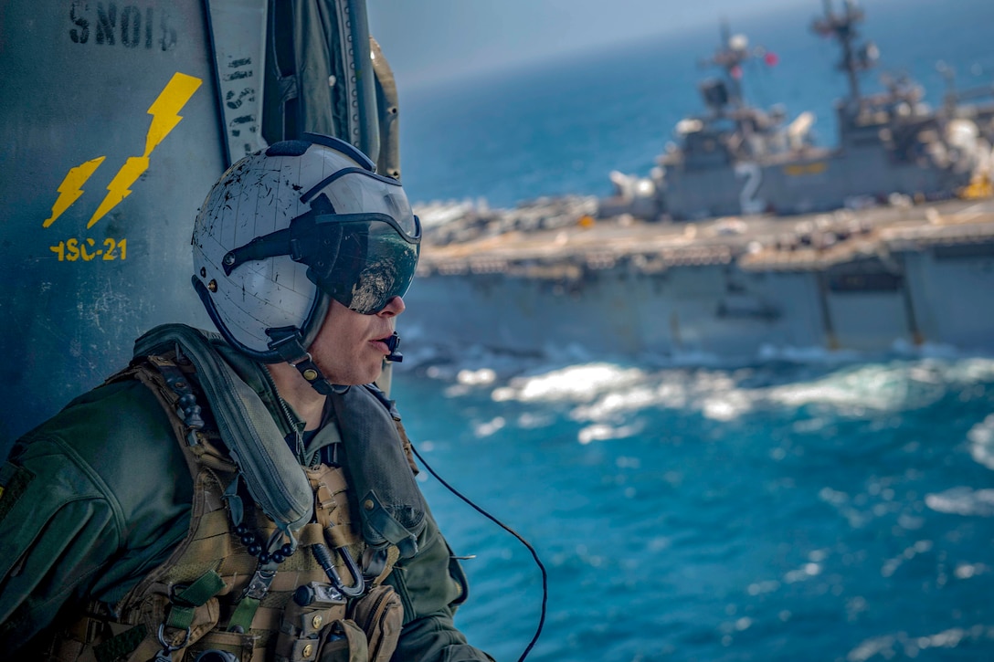 A sailor looks out over blue water and a passing ship while aboard a helicopter.