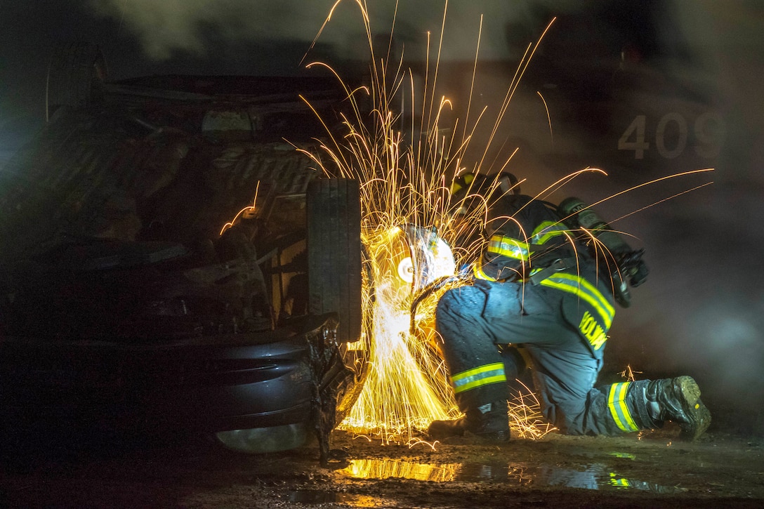 Sparks fly in the night as a kneeling soldier uses a saw to open a door on a flipped car.