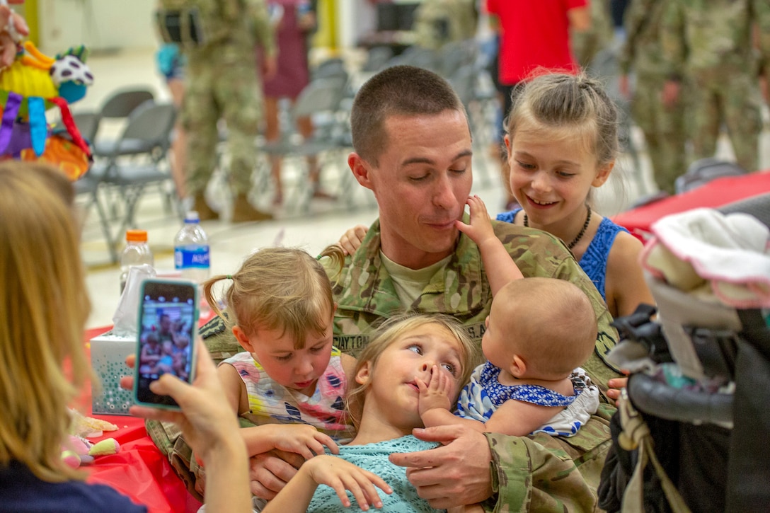 A soldier sits on a floor and holds three young children in his lap, as another stands by and a woman captures the image with her phone.