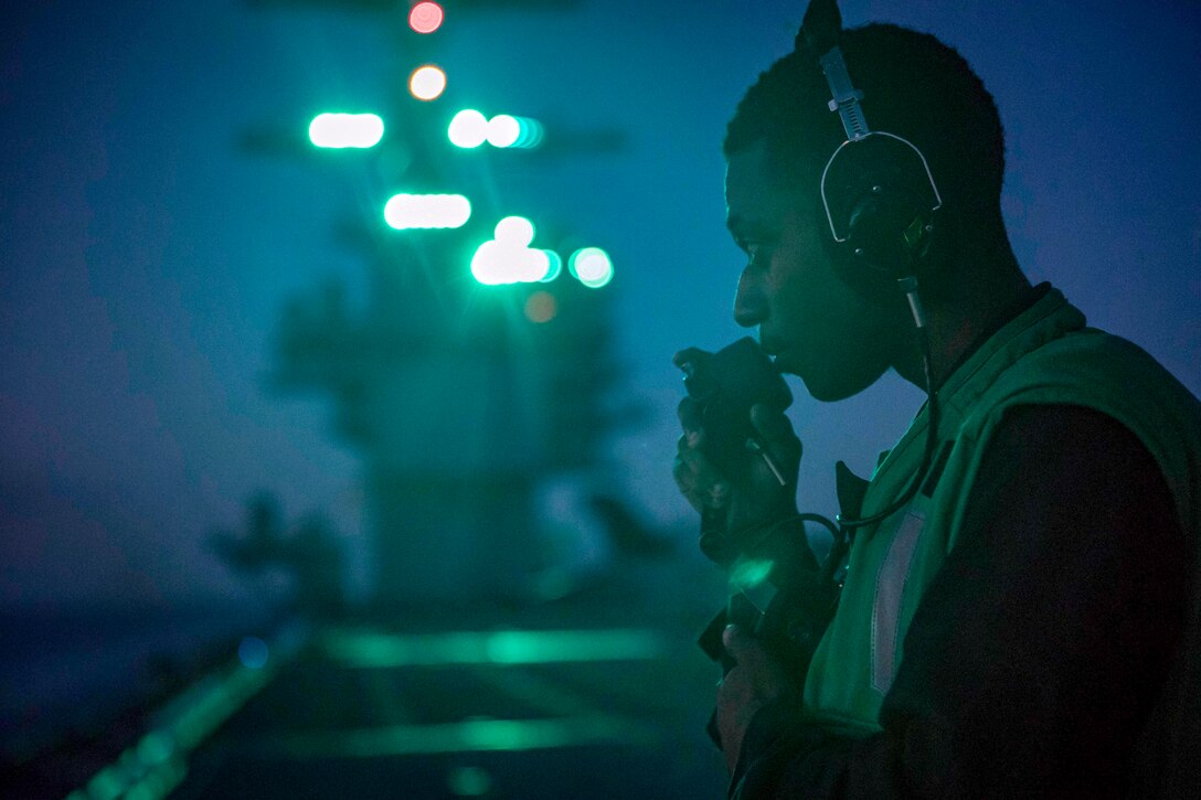A sailor wearing headphones speaks into a device on a ship's deck, against a dark blue sky.