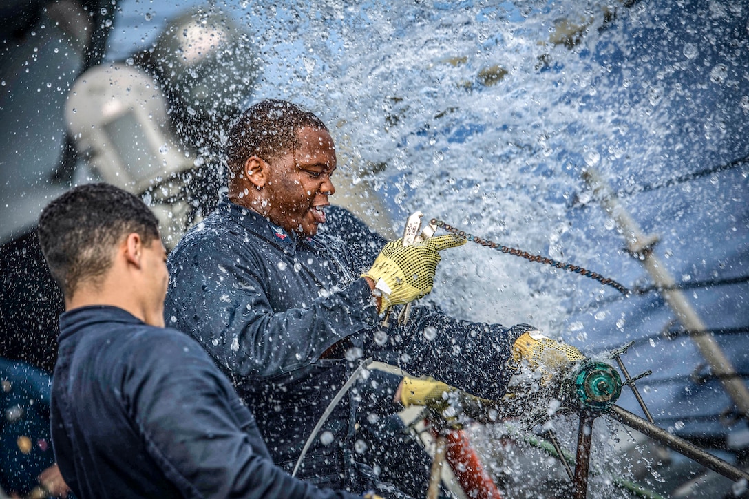 Water spews from a ruptured pipe a sailor is trying to fix, as another looks on.