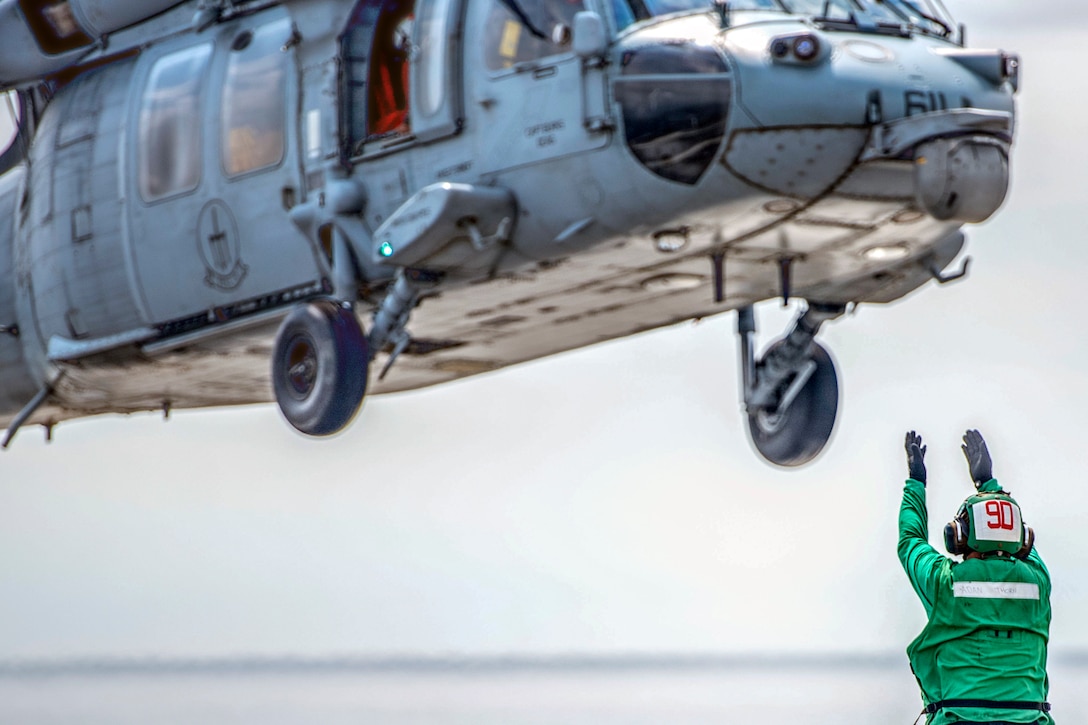 A sailor in green holds his arms up as a helicopter launches above him