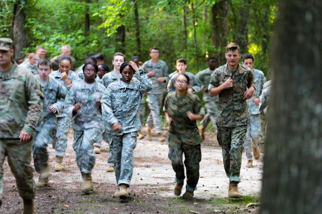 Army drill sergeants and Junior Reserve Officers’ Training Corps cadets run through a forest.