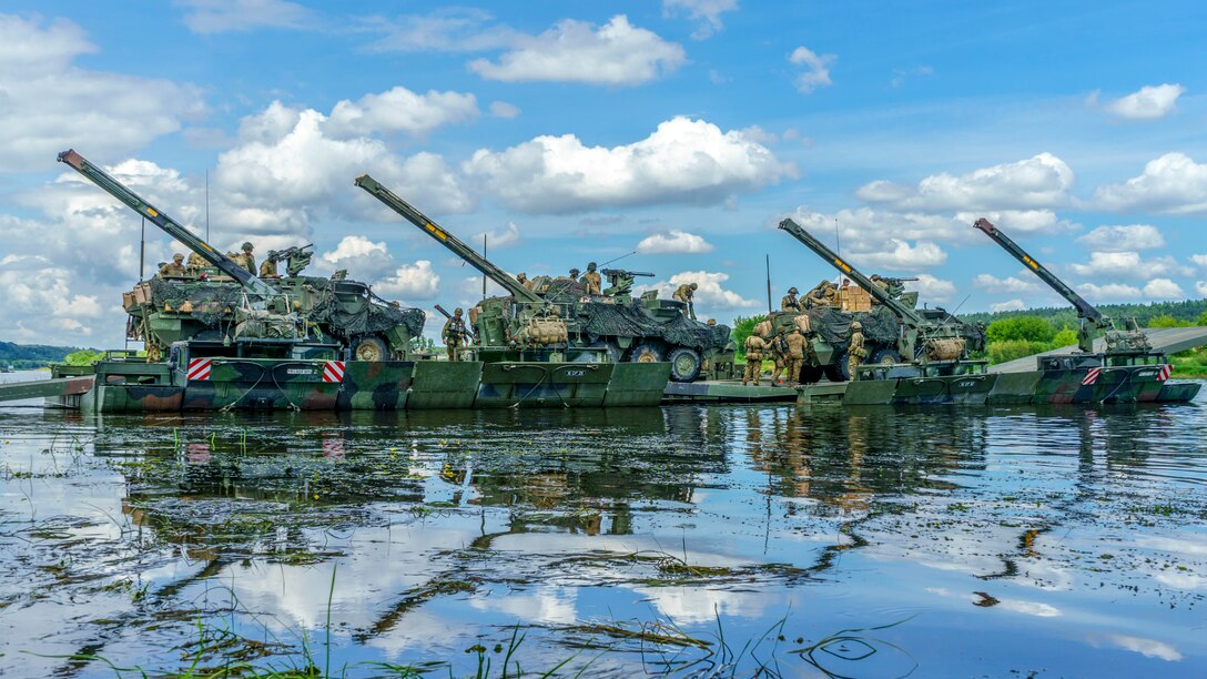 Tanks and service members cross a river on an amphibious platform.