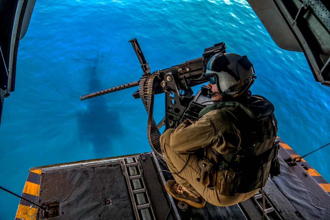 A Marine looks out from an open helicopter door over turquoise waters.
