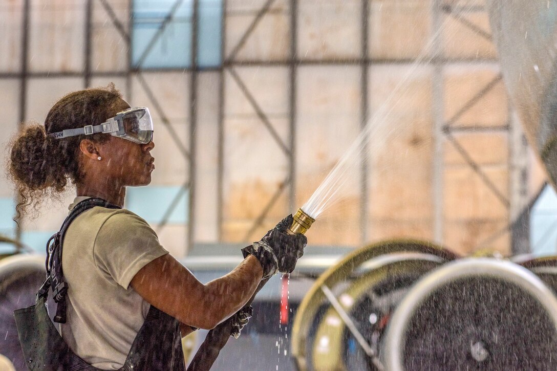 An airman wearing goggles uses a hose on the side of an aircraft in a hangar.