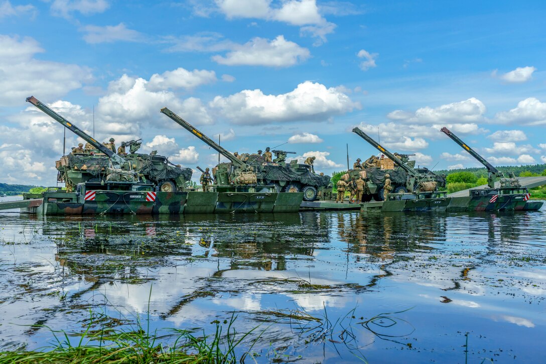 Tanks and service members cross a river on an amphibious platform.