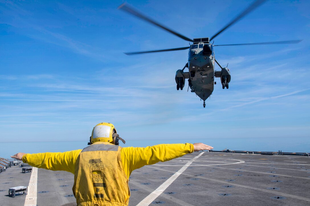 A sailor uses hand signals directing a German helicopter as it lands on the flight deck.