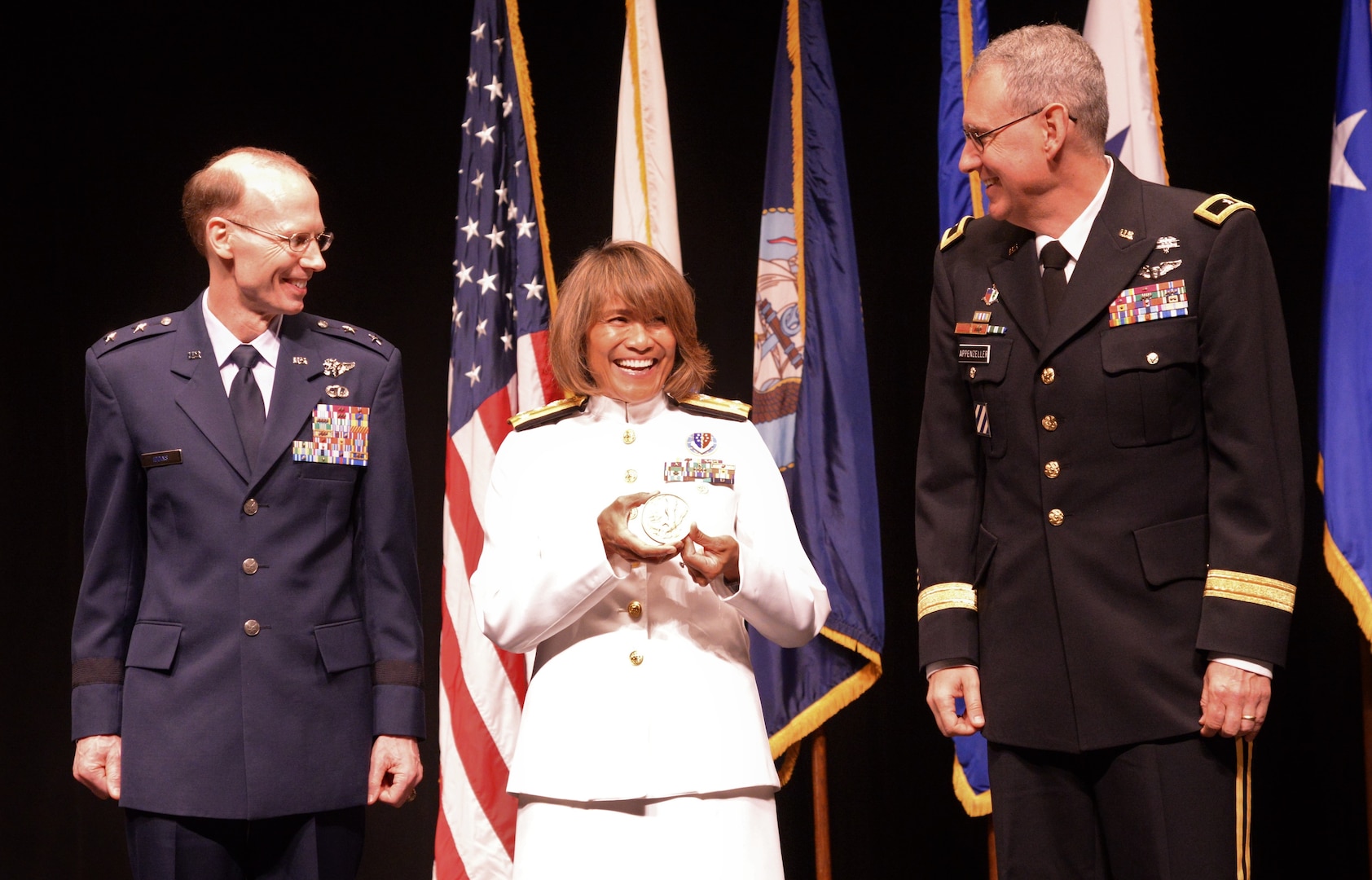Air Force Maj. Gen. Bart O. Iddins (left), 59th Medical Wing commander, and Army Brig. Gen. George Appenzeller (right) , Brooke Army Medical Center commander, present keynote speaker Vice Adm. Raquel Bono, director of the Defense Health Agency, with a US Navy Bicentennial Bronze medal during the San Antonio Uniformed Services Health Education Consortium graduation ceremony held June 7 at the Lila Cockrell Theatre in downtown San Antonio.  The front side of the coin is a Surface ship, aircraft carrier, fighter jet and submarine, while the reverse has the Flagship Alfred of the First American Fleet serving from 1775-1778 and the American eagle grasping an anchor.
