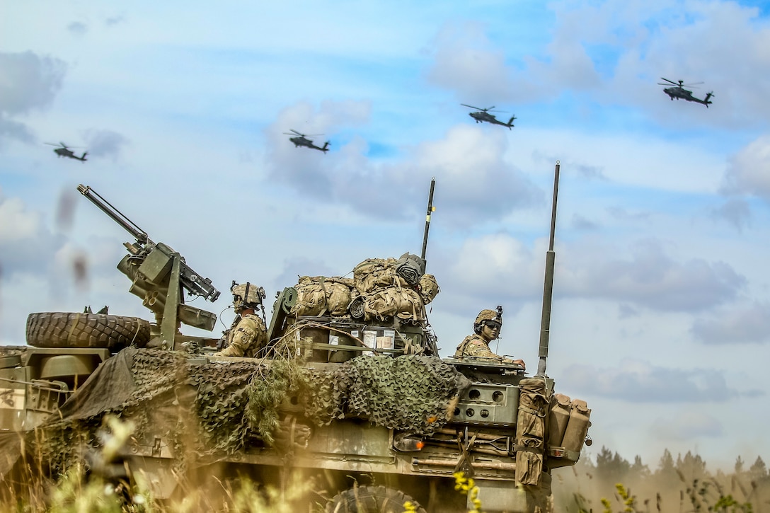 Helicopters fly over an armored vehicle.