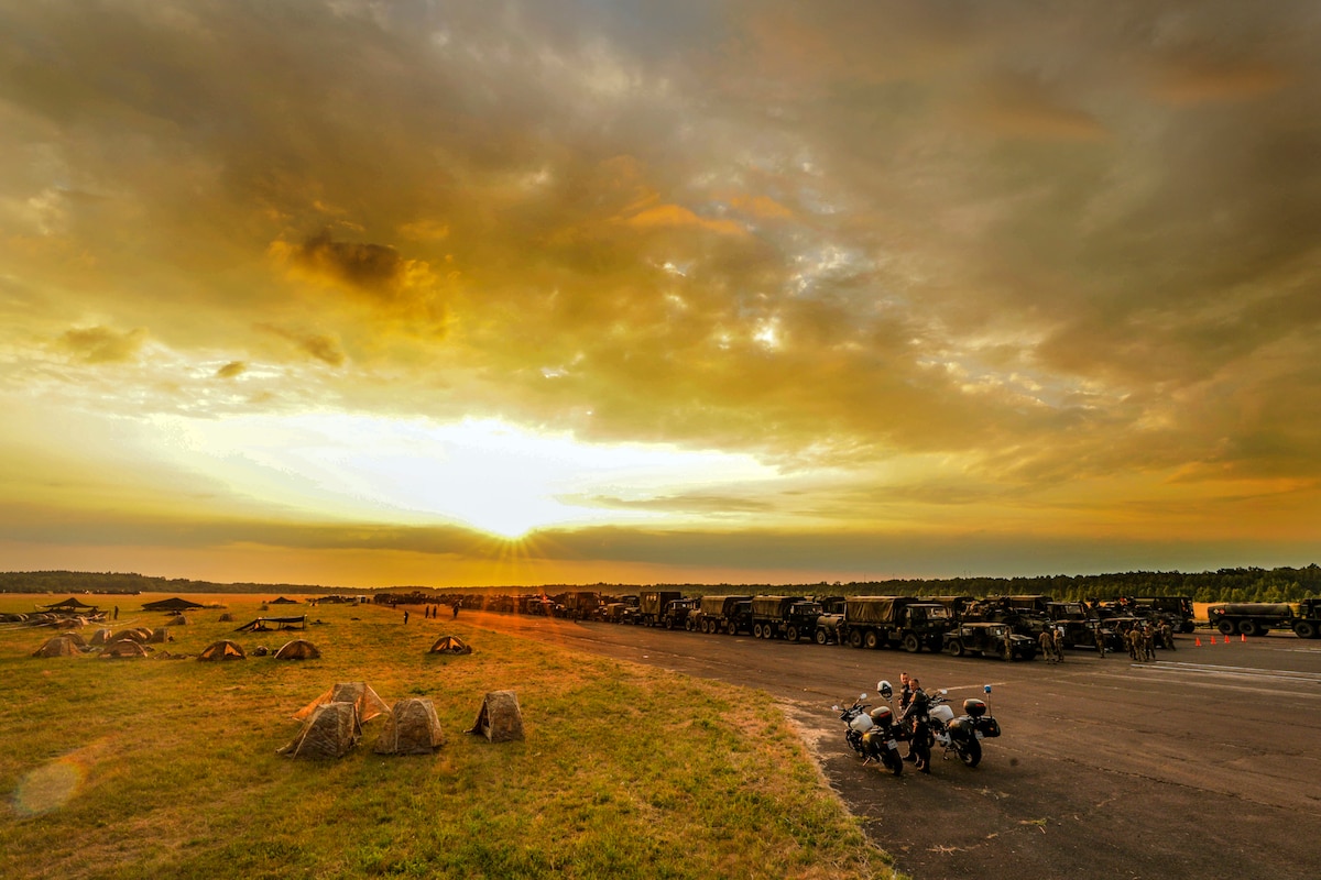 Vehicles sit parked in rows in a field, with a sun-streaked orange-yellow sky above.