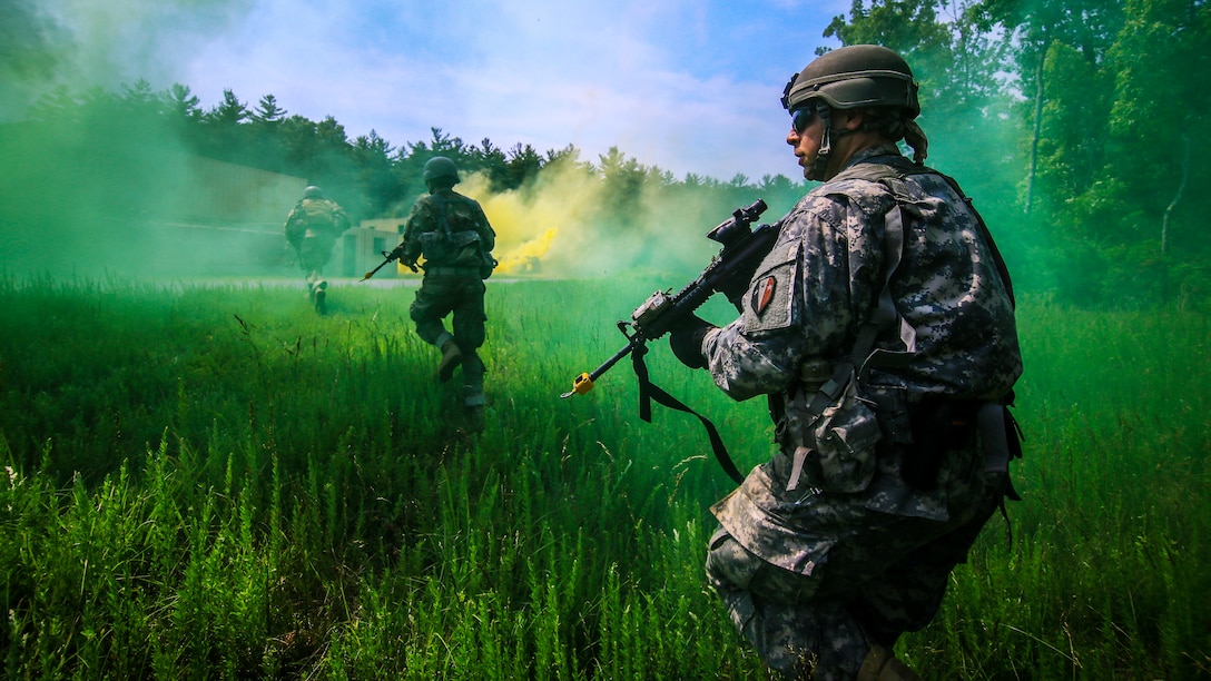 A soldier in the background runs across a field toward green and yellow smoke, as another one kneels in the foreground.