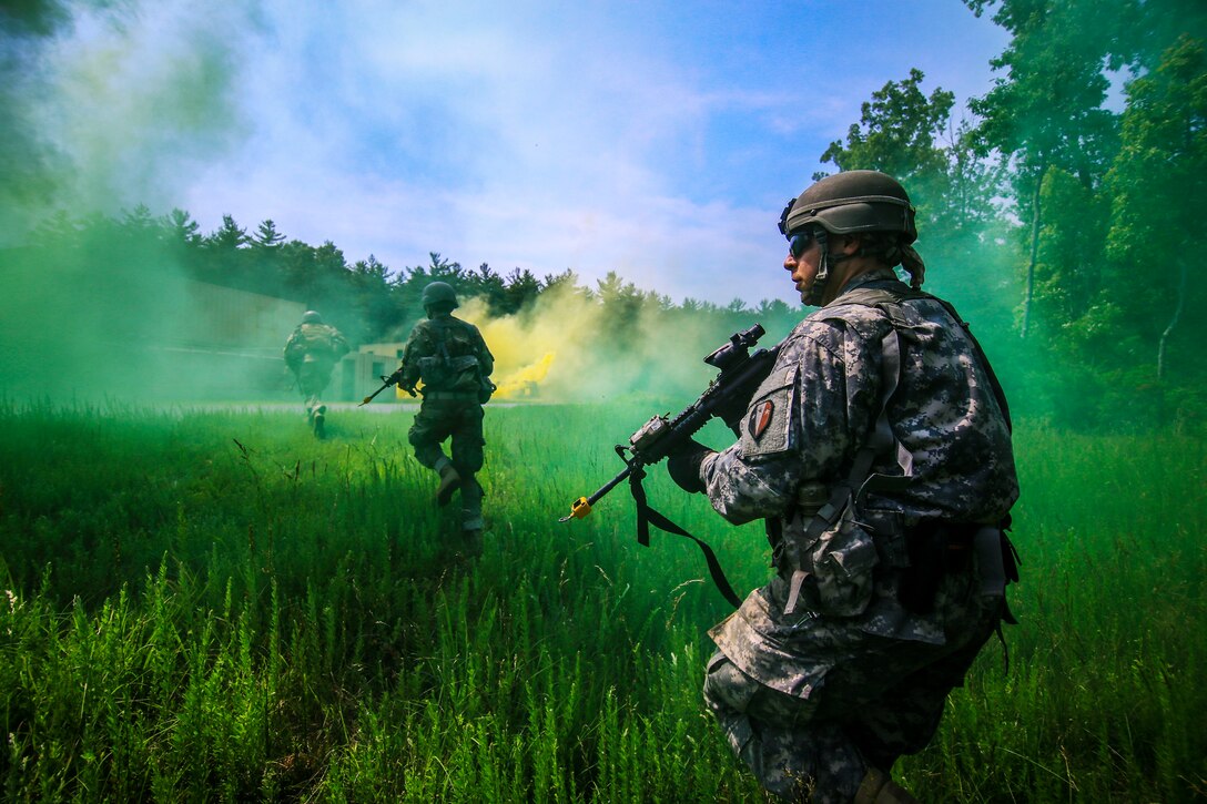 A soldier in the background runs across a field toward green and yellow smoke, as another one kneels in the foreground.