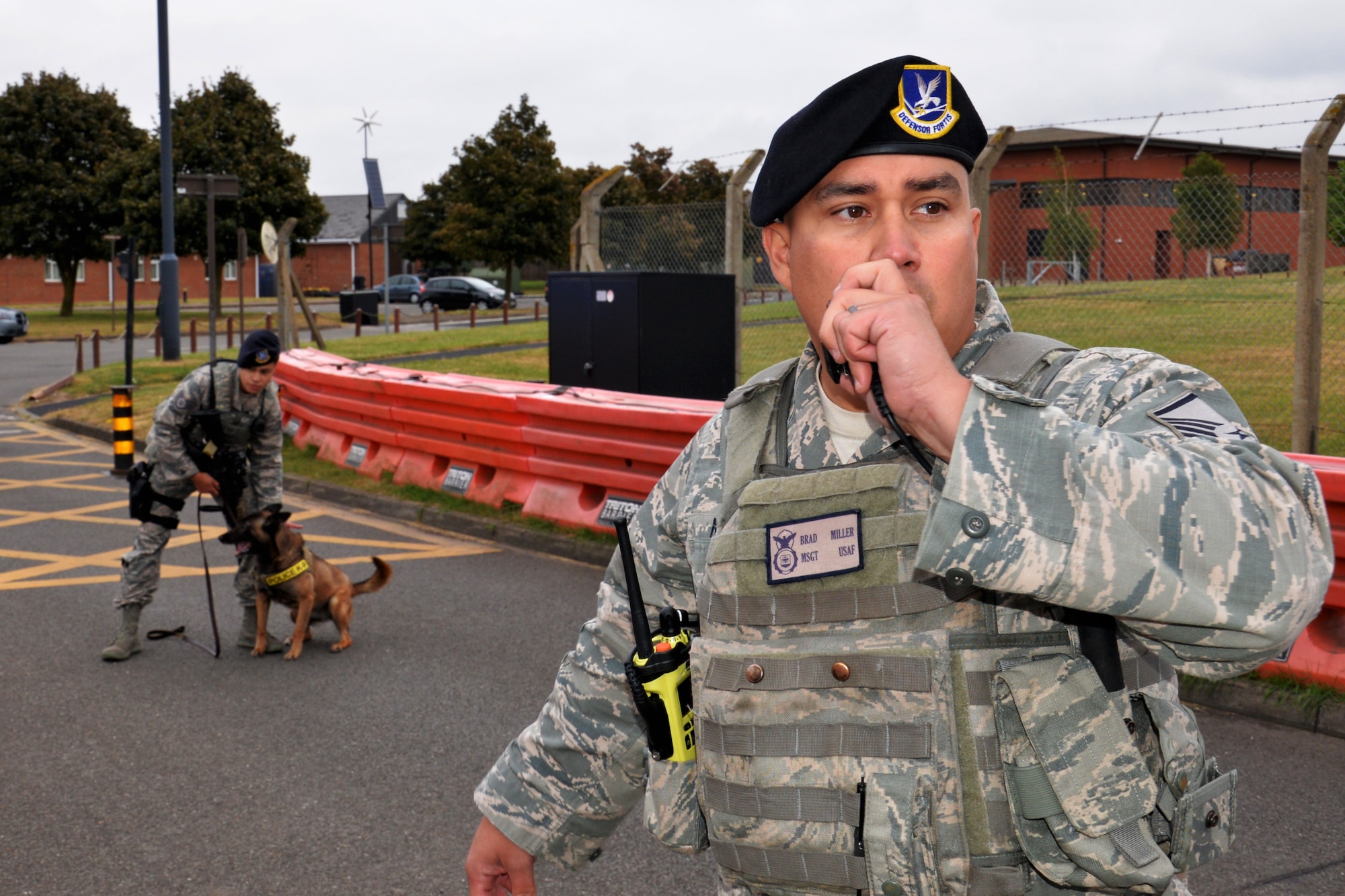 U.S. Air Force Master Sgt. Brad Miller, 100th Security Forces Squadron flight chief, speaks into his radio while Staff Sgt. Kristina Santos and Military Working Dog Ukkie stand ready in response to a simulated protest incident during an exercise at RAF Mildenhall, England, June 20, 2018. Such exercises are regularly scheduled to test responsiveness and readiness of base personnel. (U.S. Air Force photo by Tech. Sgt. David Dobrydney)