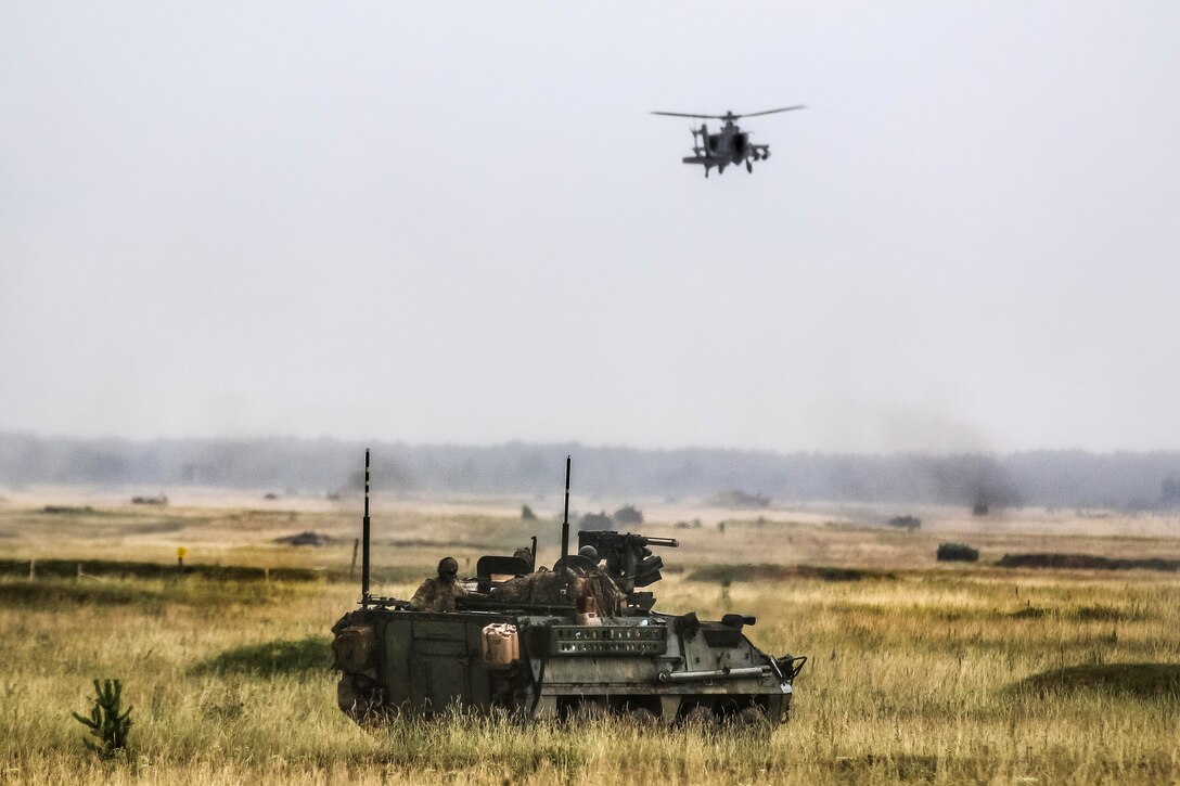 An Army M1126 Stryker and an AH-64 Apache helicopter move forward to secure an area.
