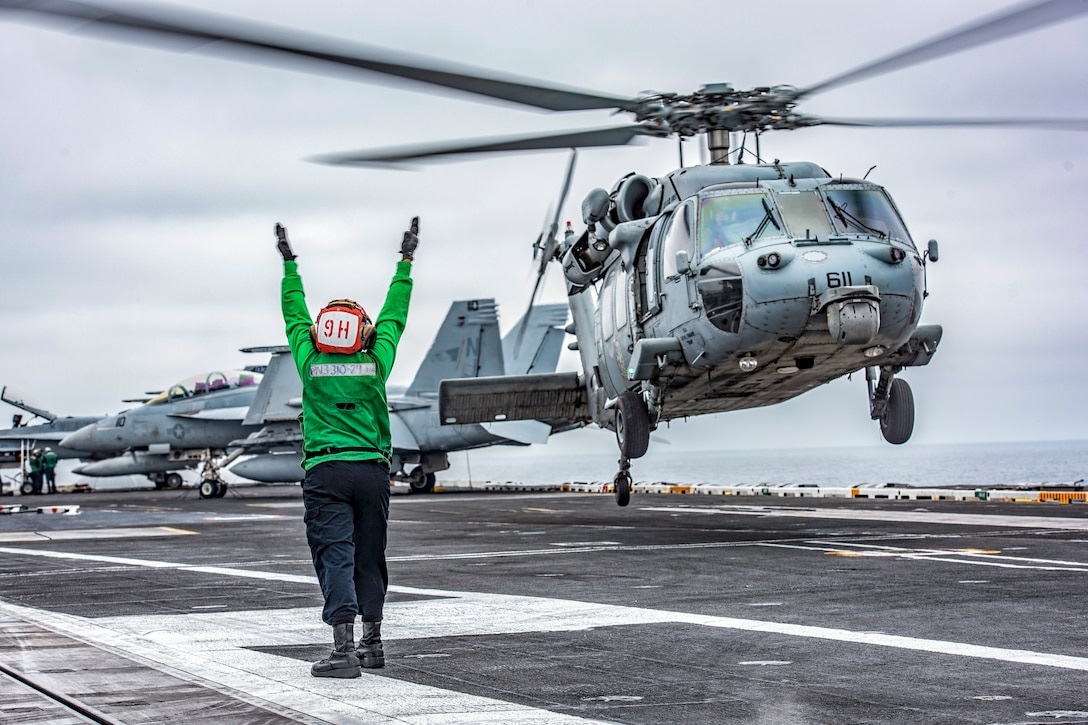 A sailor in green, shown from behind, extends her arms upward as a helicopter takes off from a ship's flight deck.