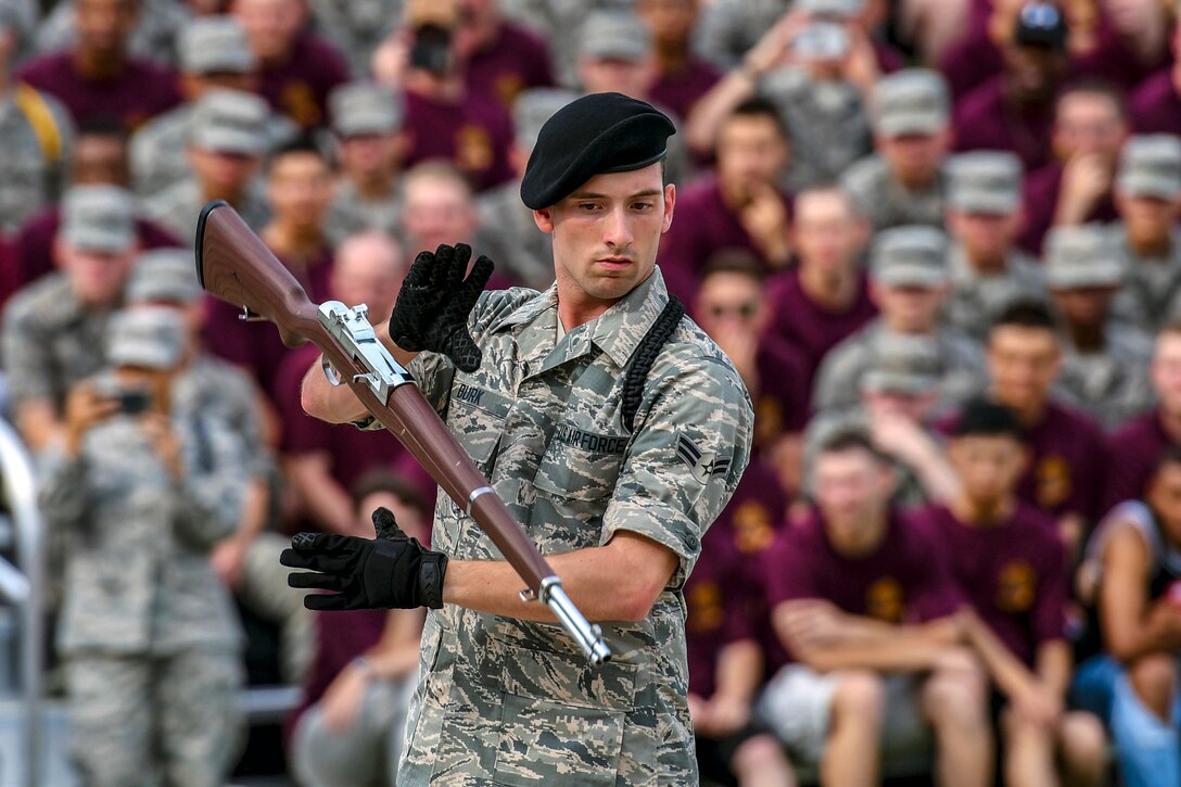 An airman in black gloves performs a drill routine with a rifle as a crowd watches.