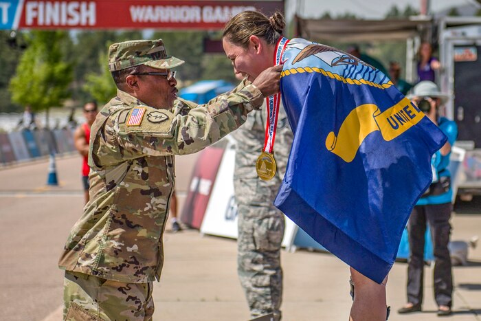 A service member puts a gold medal around the neck of another service member, who is wearing a flag.