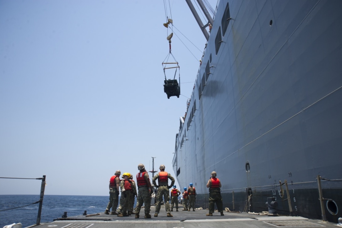 Sailors and soldiers lower an ambulance to a ferry's flight deck using cranes.