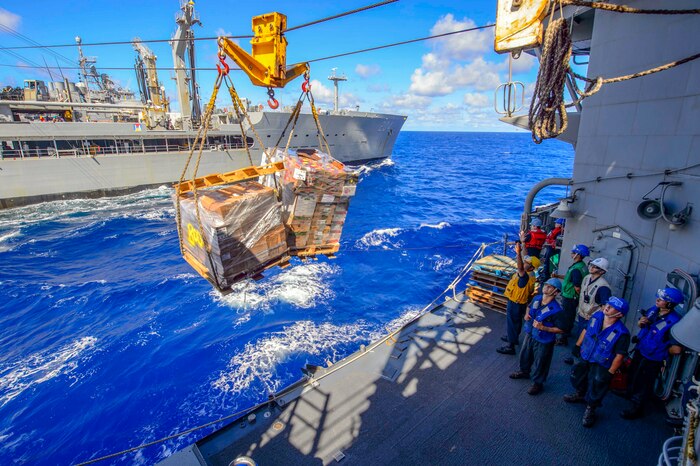 Sailors stand on a ship and watch cargo suspended on a line.