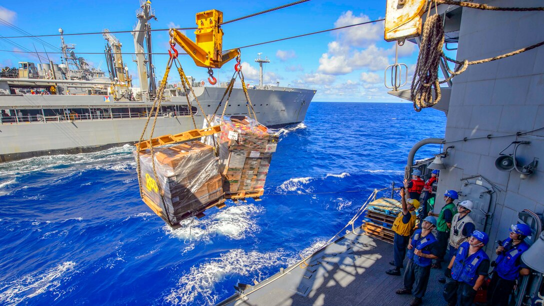 Sailors stand on a ship and watch cargo suspended on a line.