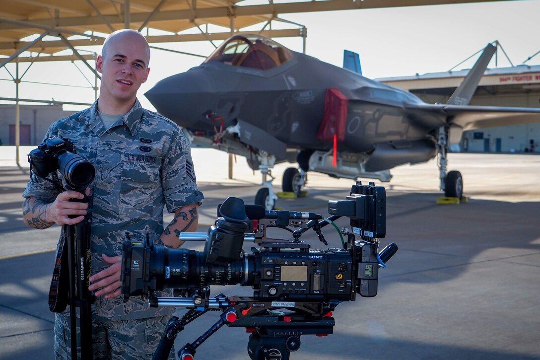 An Air Force reservist stands with a camera on a tripod in front of a military jet.