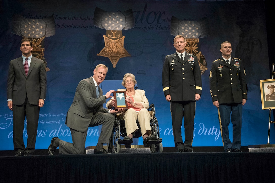 Deputy Defense Secretary Patrick M. Shanahan kneels next to a woman in a wheelchair on a stage as they hold up a framed flag.
