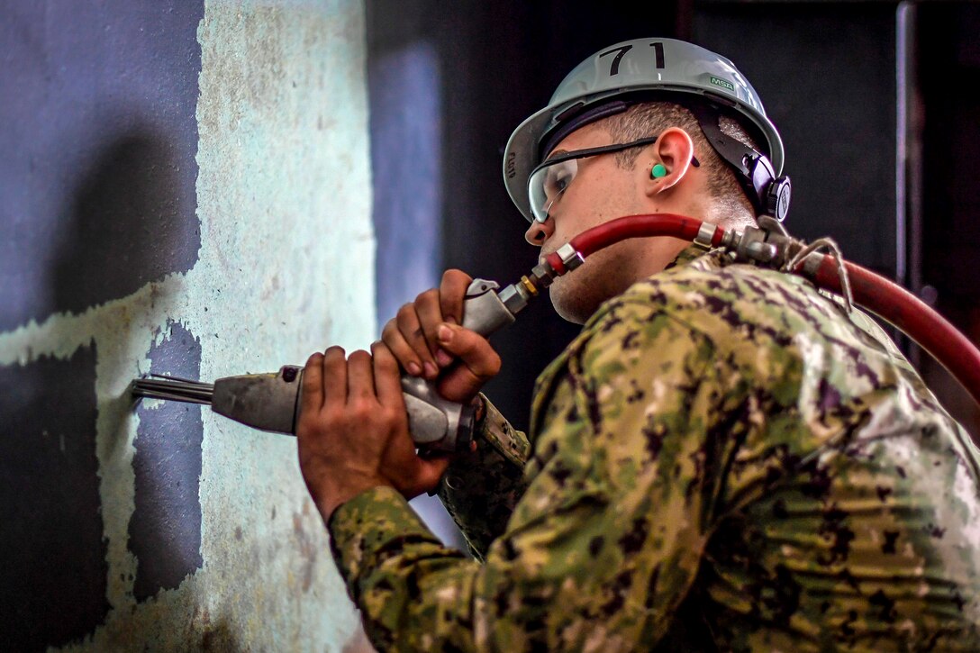 A sailor uses a power tool to chip paint off a wall.