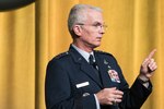 Air Force Gen. Paul J. Selva holds up an index finger while speaking in front of a mustard-colored curtain.