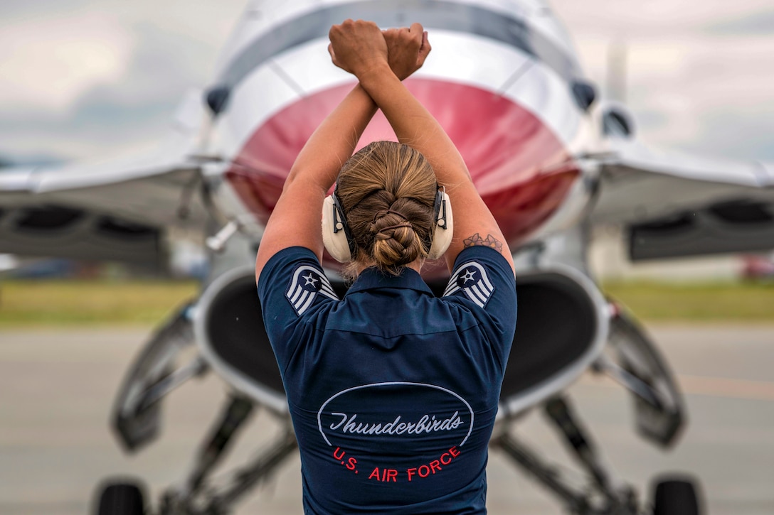 An airman, shown from behind, crosses her arms over her head in front of a jet's nose.