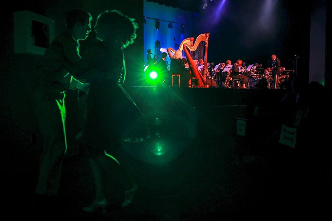 Swing dancers, illuminated in green light, dance in the foreground as as Army musicians perform on stage in the background.