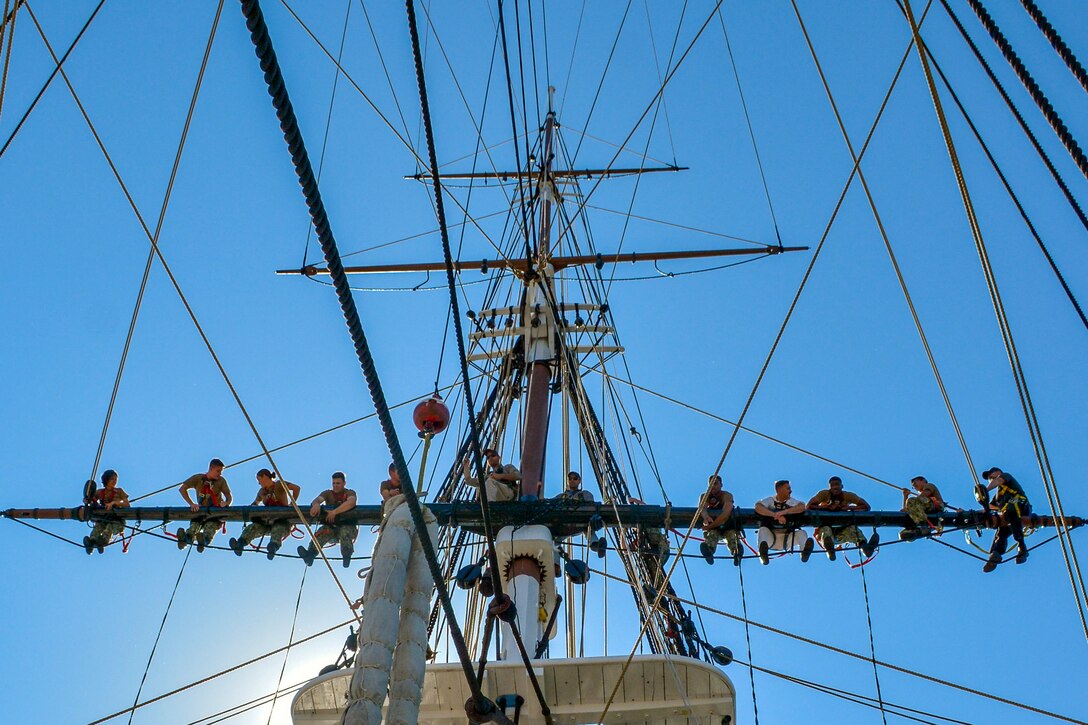 Sailors sit in a row on a ship's mast.