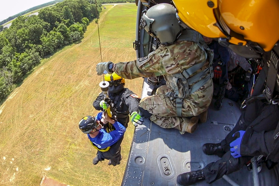 A soldier in a helicopter holds onto a hoist lifting up two people over brown terrain.