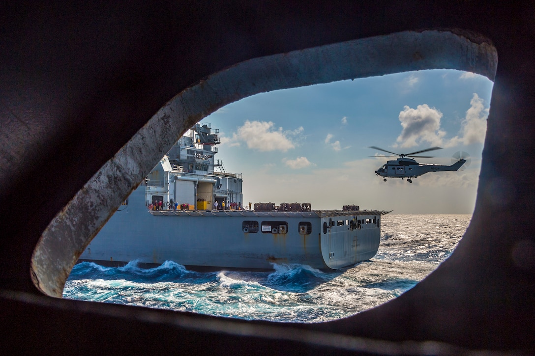 A ship's port hole shows a helicopter flying near another ship in the ocean.