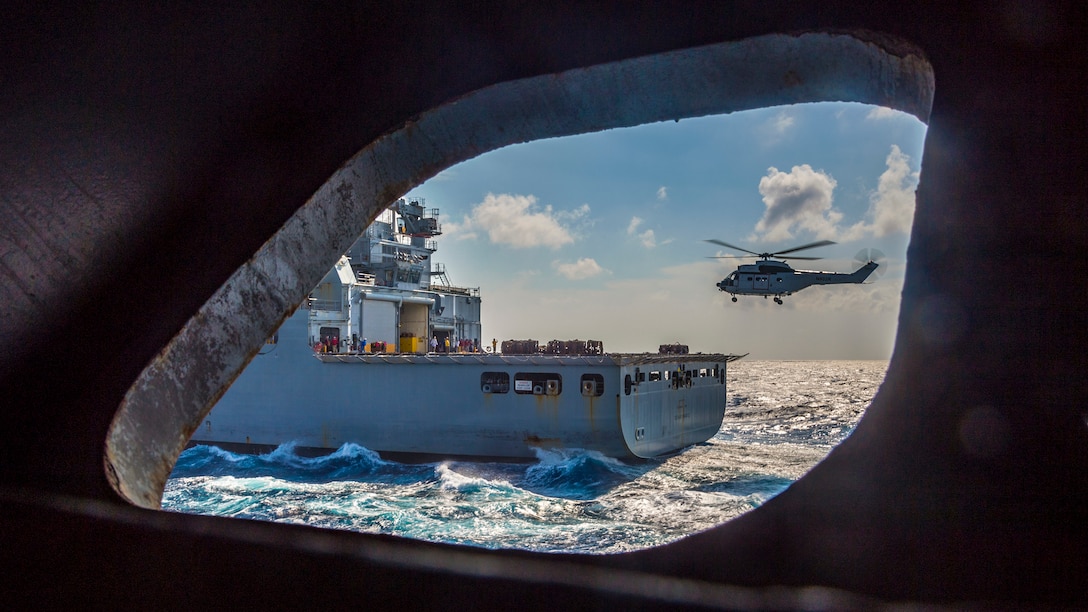 A ship's port hole shows a helicopter flying near another ship in the ocean.