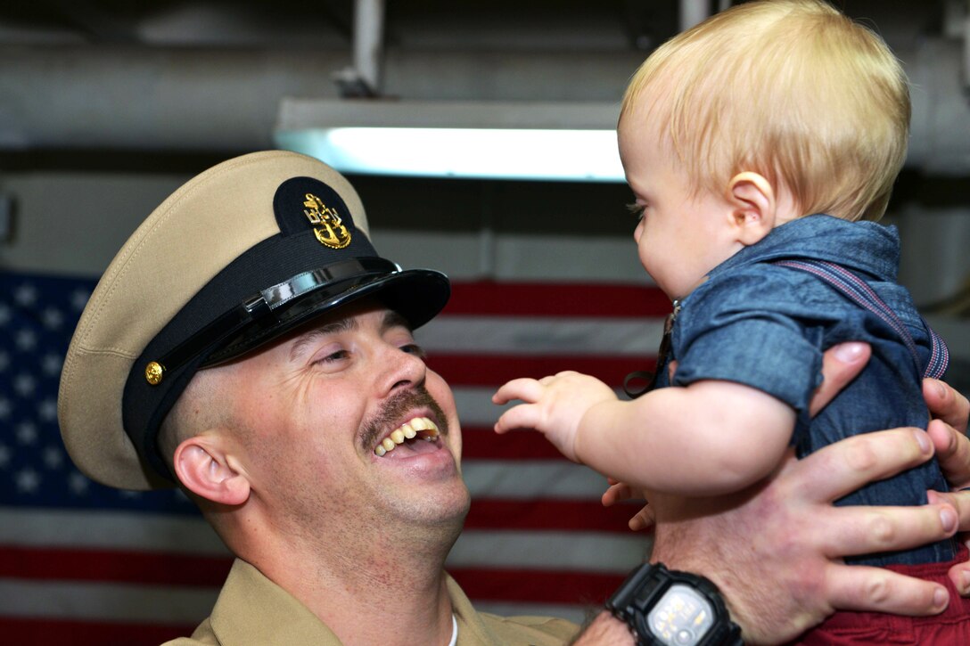 A sailor holds a child with an American flag behind him.